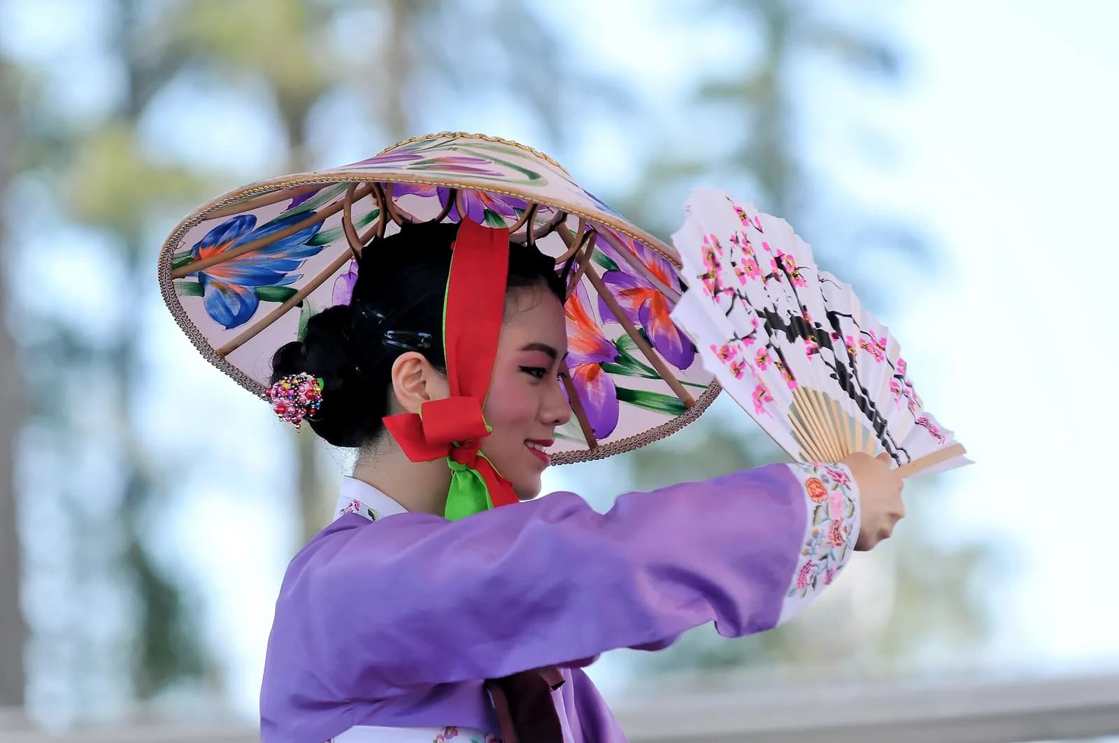 A woman in a purple dress and a large, colorful hat holds a white fan with cherry blossom designs.