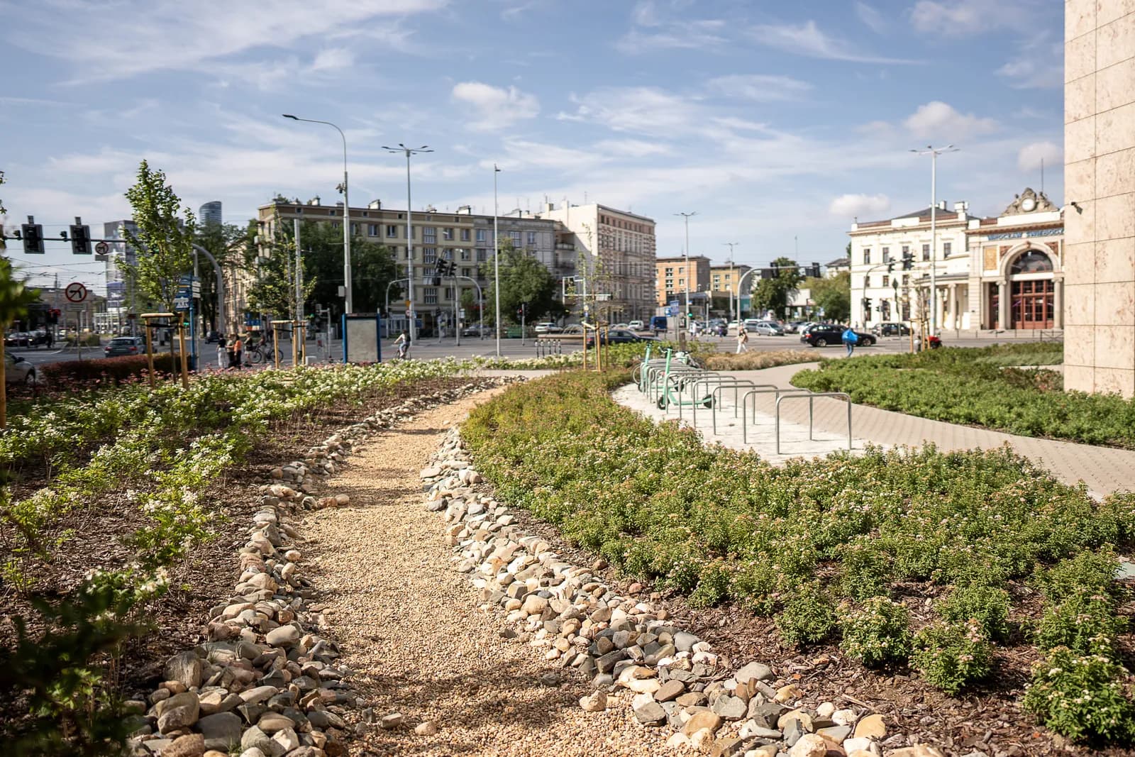 A landscaped city square with a gravel path bordered by rocks and green plants, leading to a street with cars, pedestrians, and historic buildings under a cloudy sky.