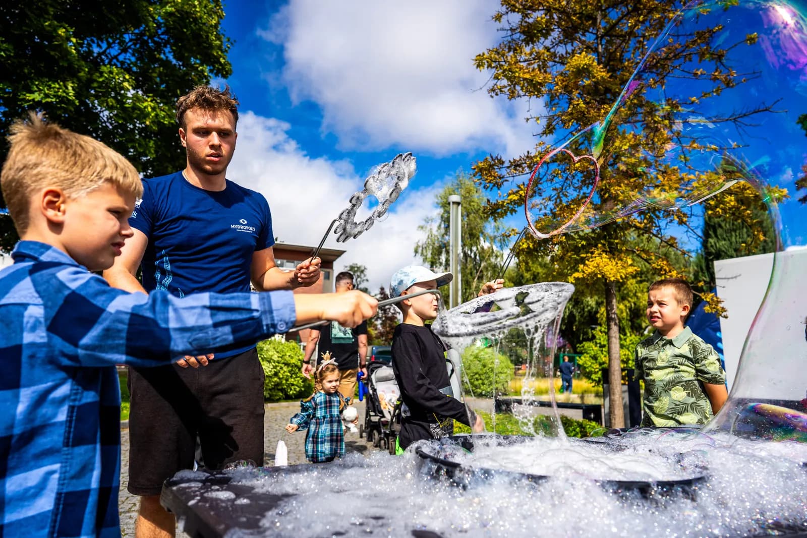 A man and several children are outdoors, creating large bubbles with wands and a soapy solution in a black container.